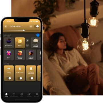A person relaxing on a sofa in a living room with warm hanging bulbs above and a smartphone app controlling the lighting scenes.