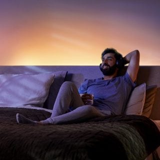Person relaxing on a bed in a bedroom, listening to music with headphones, ambient backlighting enhances the cozy setting.