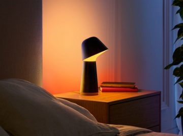 A modern table lamp with a dark base emits warm light on a bedside table, illuminating nearby books and the wall behind it.