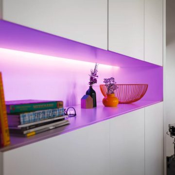 LED light strip installed under a white shelf casts purple light on books, vases, and a wire bowl placed on the shelf.