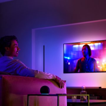 A person sits on a sofa in a living room watching TV, with colorful ambient lighting illuminating the space.
