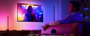 Person watching TV in a living room illuminated by smart colored lights, with a voice assistant device on the table.