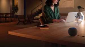 A person works on a laptop at a wooden table in a softly lit living space, with a smart speaker and books nearby.