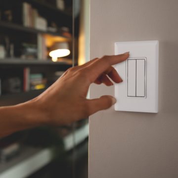 Hand pressing a white smart light switch on a wall, illustrating control of lighting, with shelves and lamp blurred in background.