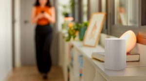 A person walks in a hallway with a white smart speaker and a round smart light on a shelf, illuminated by soft colored lighting.