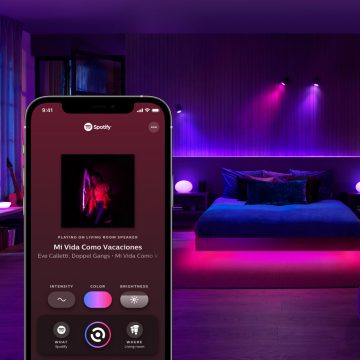 A smartphone displays a Spotify interface in front of a bedroom with smart LED lighting casting purple and pink light on walls and furniture.