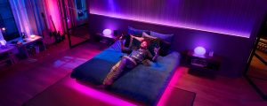 Person relaxing on a bed in a bedroom, surrounded by colorful smart lighting in shades of pink, purple, and blue.