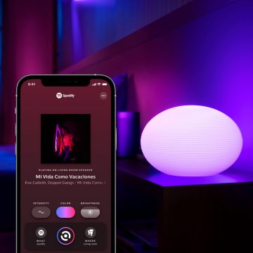 A smartphone displays a Spotify music sync app with intensity, color, and brightness controls beside a glowing oval smart lamp.