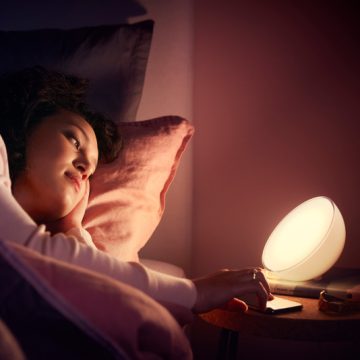 Person in bed using a portable LED lamp on a bedside table, warm light illuminating the bedroom setting.