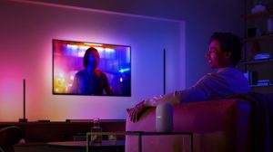 Person watching TV in a living room with smart lighting casting colorful hues on walls and furniture, voice assistant device visible.