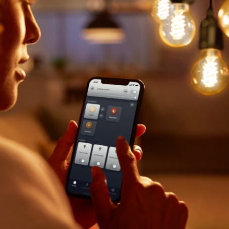 Person adjusting lighting settings on a smartphone in a warmly lit room with vintage-style hanging bulbs in focus.