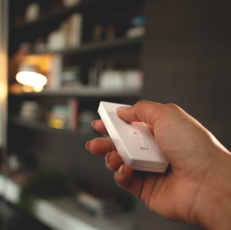 Hand pressing a button on a white smart lighting remote control, with a blurred bookshelf and lamp in the background.