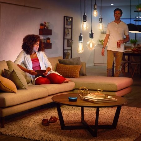 Two people in a living room, one seated on a sofa using a tablet, with warm filament light fixtures illuminating the space.