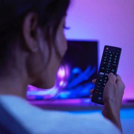Person holding a TV remote in a living room, illuminated by purple and blue ambient lighting from the television screen.