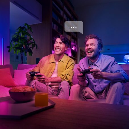 Two people playing video games on a sofa in a living room, illuminated by vibrant colored smart lighting with snacks on the table.