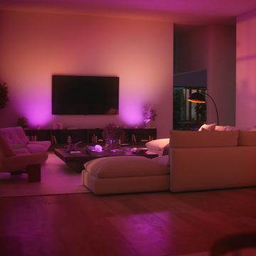 A living room with soft ambient purple and pink lighting, modern furniture, and a large screen on the wall creating a relaxing scene.