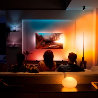 Three people sit on a sofa watching a TV with colored smart lighting matching the screen in a modern living room.