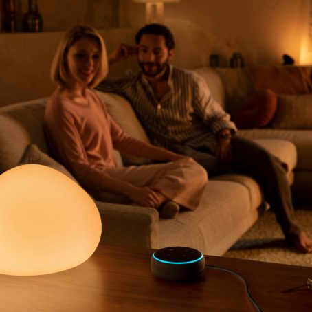 Two people sitting on a sofa in a living room, using voice control with a smart speaker and a glowing table lamp nearby.