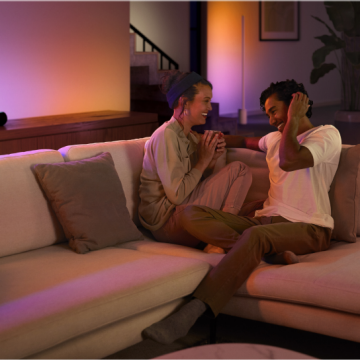 Two people sitting on a living room sofa talking, with gradient colored smart lighting illuminating the background.