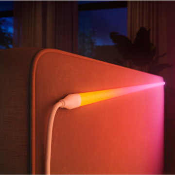 A flexible lightstrip with a white connector is attached to the back of a sofa, emitting a gradient of colored light onto the fabric.