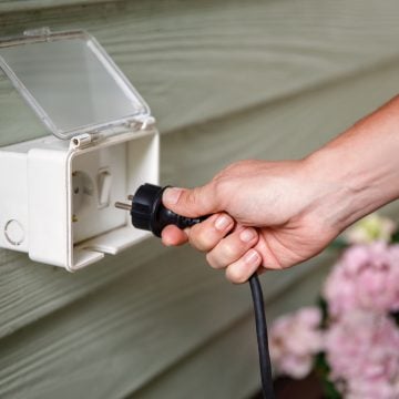 Hand plugging a black electrical cord into an outdoor socket with a protective cover, suggesting setup for outdoor lighting.