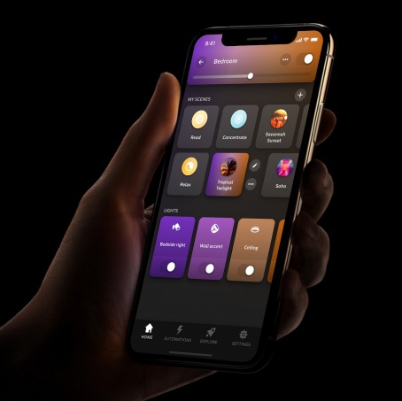 A hand holds a smartphone displaying a smart lighting app with scene options, light controls, and a brightness slider for the bedroom.