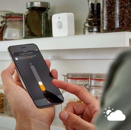 A person adjusts daylight sensitivity for a motion sensor on a smartphone in a kitchen with shelves holding jars and spices.