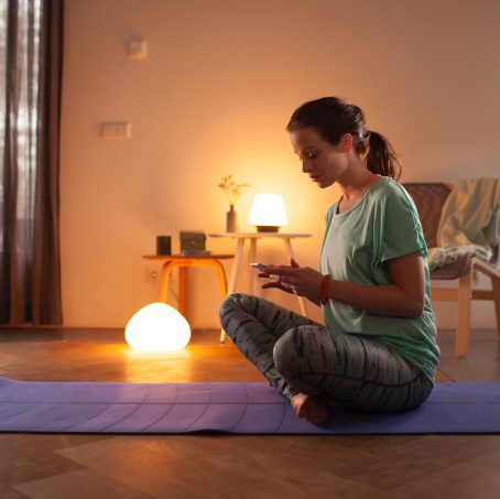 Person sitting cross-legged on a yoga mat in a living room, using a phone, with smart light kits providing soft warm illumination.