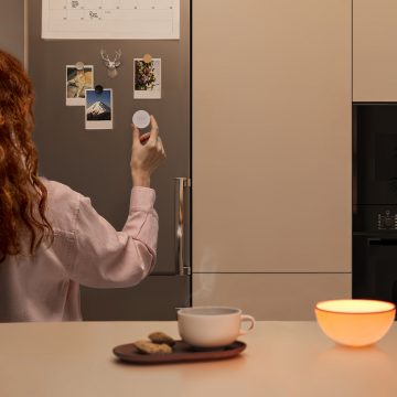 Person in a kitchen uses a round smart switch on a fridge, with a glowing LED light and cup of tea on the counter.
