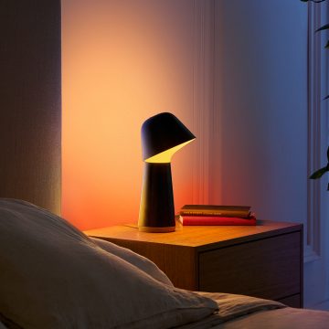 A modern smart table lamp with a dark finish placed on a bedside table, casting LED light onto the wall and surface.