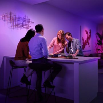A group of people gather around a kitchen island sharing food, illuminated by colorful smart lighting in a modern dining area.