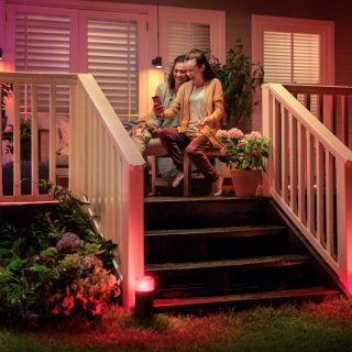 Two people sitting on a porch using a smartphone, with smart outdoor lights illuminating the steps and garden area.