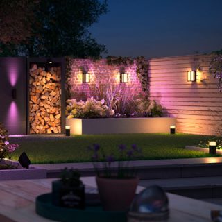 Outdoor garden lights mounted on walls and placed along pathways, illuminating wood storage, plants, and fence with colored light.