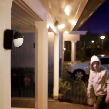 A motion sensor is mounted on an outdoor wall, activating porch lighting as a person in a hoodie approaches a driveway at night.