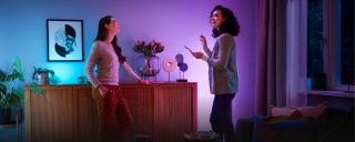 Two people conversing in a living room, illuminated by colorful smart lighting on a sideboard with decorative objects and plants.