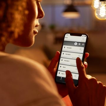 A person adjusts indoor smart lighting with a mobile app in a warmly lit living space, with visible hanging light bulbs.