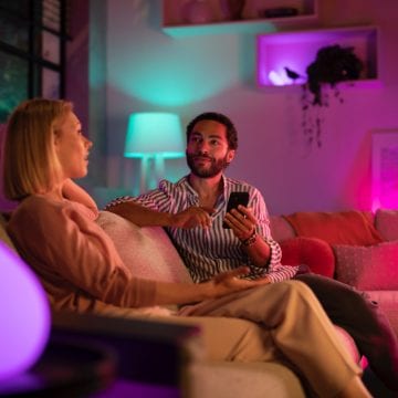 Two people sitting on a sofa in a living room, using a smartphone, with colorful indoor ambiance lighting illuminating the space.