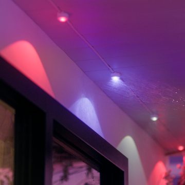 A collection of permanent string lights is mounted along a ceiling outdoors, casting colored light onto the wall and ceiling surface.