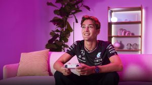 Person playing video games on a sofa in a living room with smart lighting casting purple tones on walls and shelves.