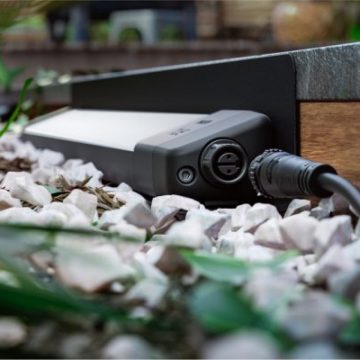 Rectangular outdoor smart light fixture placed on gravel beside a wooden edge, connected by a visible cable for exterior lighting.