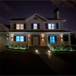 Outdoor smart lighting fixtures mounted on house facade and in garden beds, illuminating entrance, walkway, and surrounding plants at night.