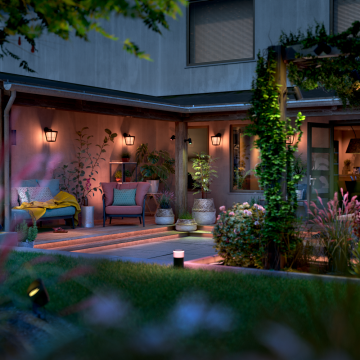 Outdoor smart lighting fixtures mounted on a backyard patio wall and garden path, illuminating seating and plants.