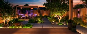 Outdoor smart lights installed along garden paths and mounted on wooden fence, illuminating plants and seating area at dusk.