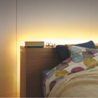 Flexible smart lightstrip installed behind a wooden headboard, emitting soft indirect light onto the wall above a bed.