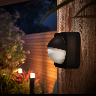 Outdoor wall-mounted smart security light with a rounded black and white design, illuminating a wooden exterior surface.