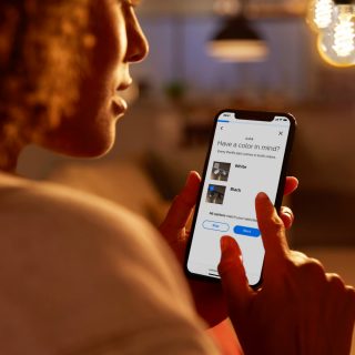 A person selects color options for track lighting on a smartphone in a warmly lit living room with pendant lights above.