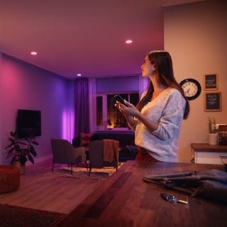 Person adjusting smart lighting with a mobile device in a modern living room with colored lights illuminating the space.