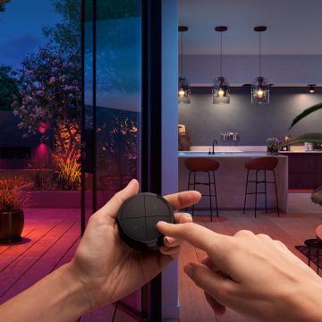 Des mains tiennent un Hue Tap dial switch, contrôlant simultanément les lampes connectées d'une cuisine et d'un jardin.