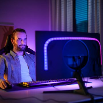 파란색과 보라색 빛을 비추는 Hue gradient 라이트스트립을 구비한 PC 모니터 앞에 앉은 한 남성. 