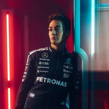 F1-coureur George Russell zit op een beige bank in een woonkamer met zachte, warme verlichting.